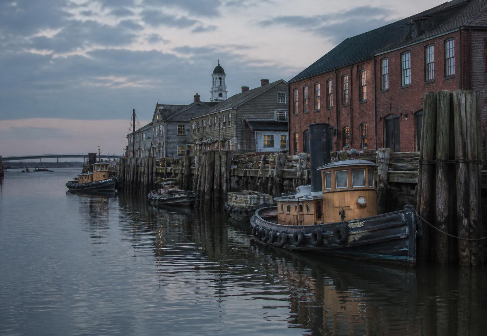 Tugboats moored beside brick warehouses on a tidal river at dusk